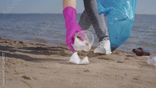 Female activist picks up plastic trash into garbage bag walking on sand of public beach on sunny day. Woman in rubber gloves collects scattered rubbish to protect environment from pollution closeup photo