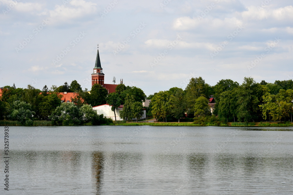 A view of a small river or lake located in the middle of a small village, with a fountain, a church and a dense forest or moor visible in the distance on a cloudy summer day in Poland
