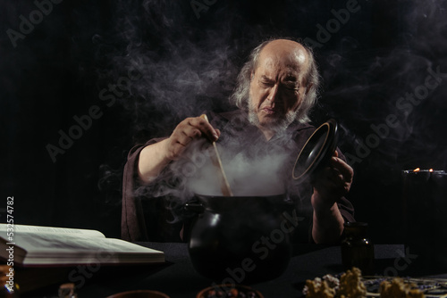 senior alchemist frowning near steaming pot while cooking at night on black background.