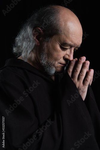 side view of senior medieval monk with praying hands near face isolated on black.