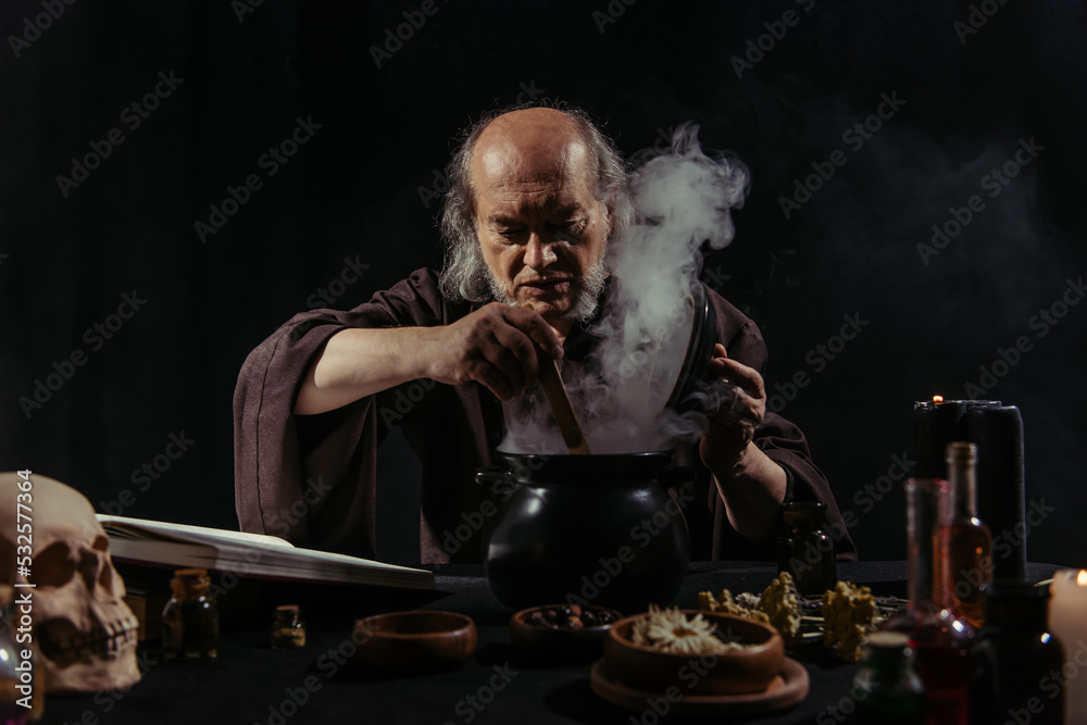 senior wizard mixing boiling potion in steaming pot at night isolated on black.