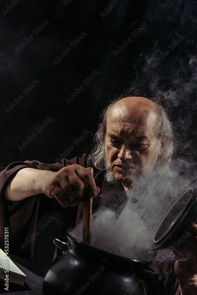 medieval healer preparing magic potion in boiling pot isolated on black.