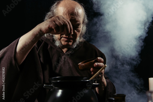 senior magician adding ingredient from bowl into pot on black background with smoke.