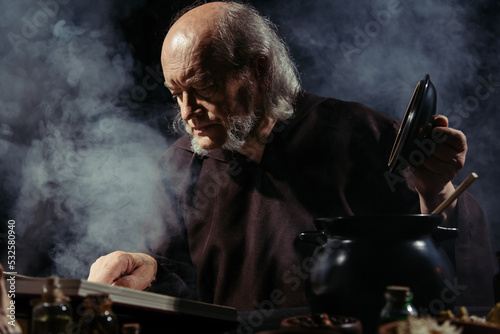 medieval alchemist reading magic cookbook while preparing potion at night on black background with smoke.