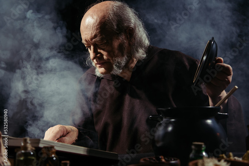 medieval alchemist reading magic cookbook while preparing potion at night on black background with smoke. photo