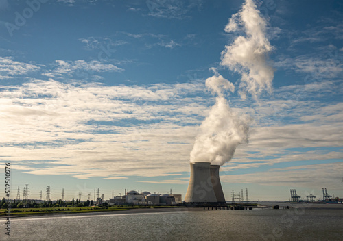 Antwerpen, Flanders, Belgium - July 10, 2022: Sunset on Cooling towers and Doel Nuclear Power plant blowing steam into blue cloudscape. Container Europa terminal on horizon photo