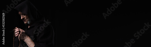 фотографія monk in dark hooded cassock praying with rosary isolated on black, banner