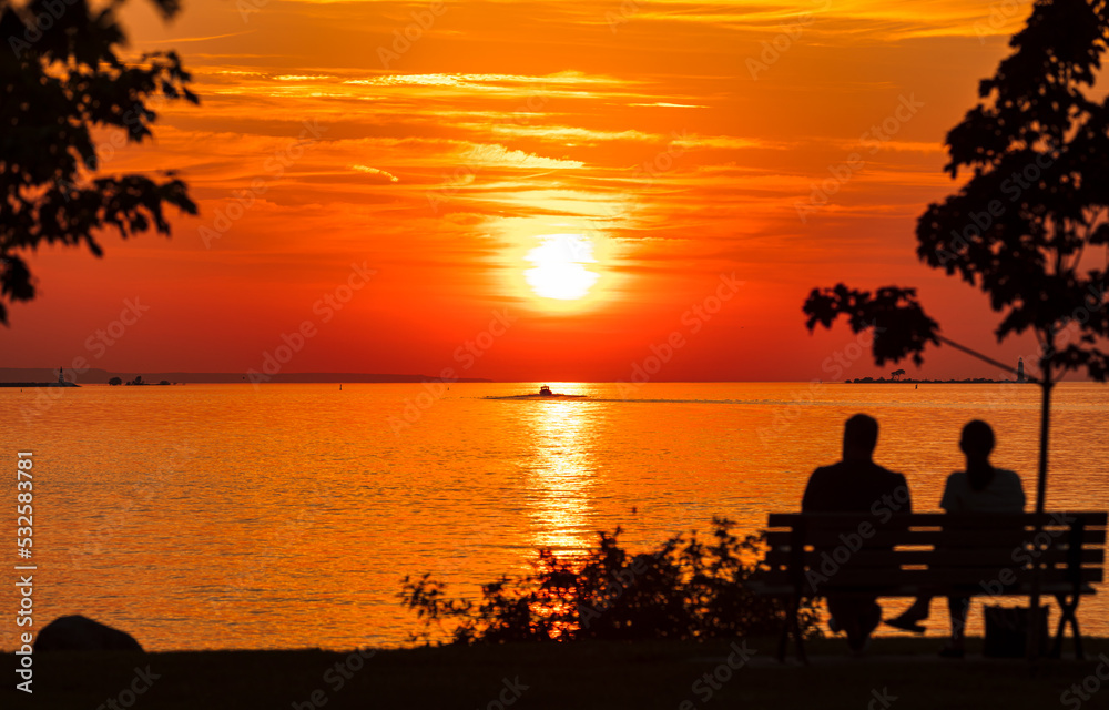 Couple sits on park bench in front of setting sun on shore of waterfront park