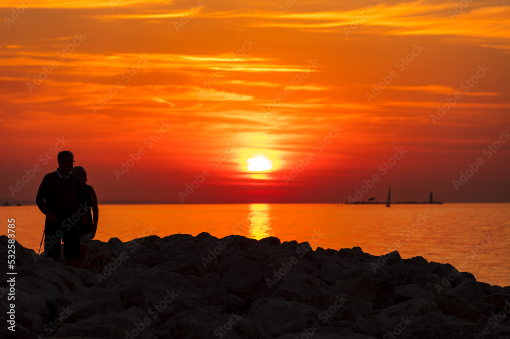 Couple stands in front of setting sun on shore of waterfront park