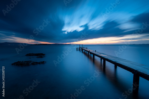 Long exposure photography of pier on the sea, dramatic clouds on skycalm sea with reflection and light on horizon