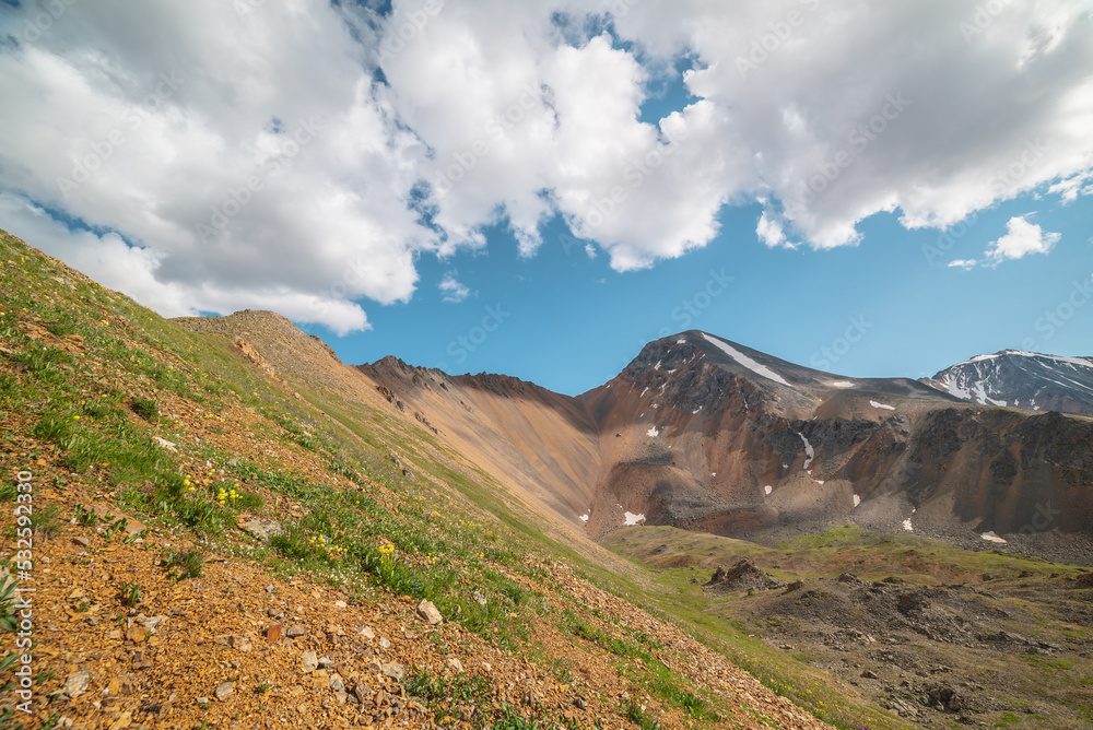 Obraz premium Scenic landscape with sunlit green grassy stone hill and high orange mountain ridge under white clouds in blue sky. Colorful scenery with rocky mountains and sharp rocks in sunlight under cloudy sky.