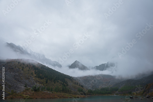 Dark atmospheric landscape with alpine lake and high mountain silhouettes in dense fog. Snowy rocky mountain top above hills in thick fog in dramatic overcast. Black rocks in low clouds during rain.