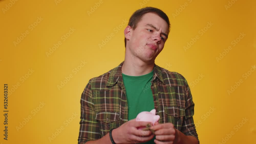 Poor man upset over insufficient amount of money, holding piggybank and one dollar banknote. Financial crisis. Bankruptcy. Poverty and destitution. Young boy isolated on yellow studio wall background Poor man upset over insufficient amount of money, holding piggybank and one dollar banknote. Financial crisis. Bankruptcy. Poverty and destitution. Young boy isolated on yellow studio wall background