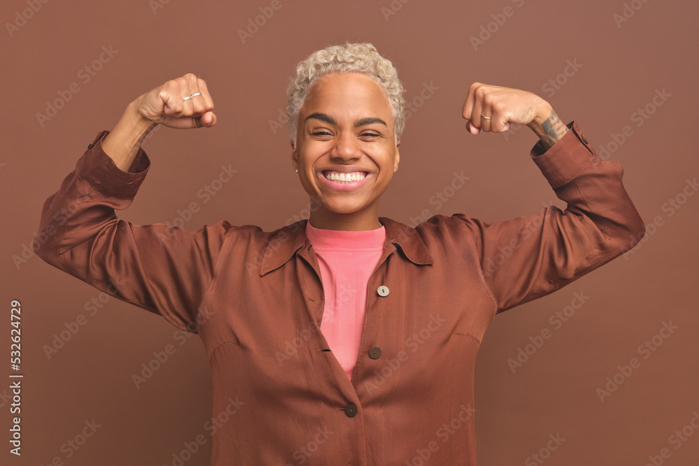 Fototapeta premium Young happy African American woman posing in Hercules pose showing off biceps as sign of self-confidence and independence or ability to achieve goals stands on plain brown background