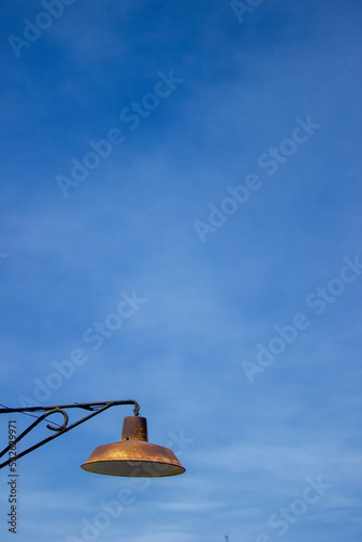 Foto vertical de un farol de metal estilo industrial antiguo oxidado y con ampolleta incandescente. De fondo el cielo azul photo