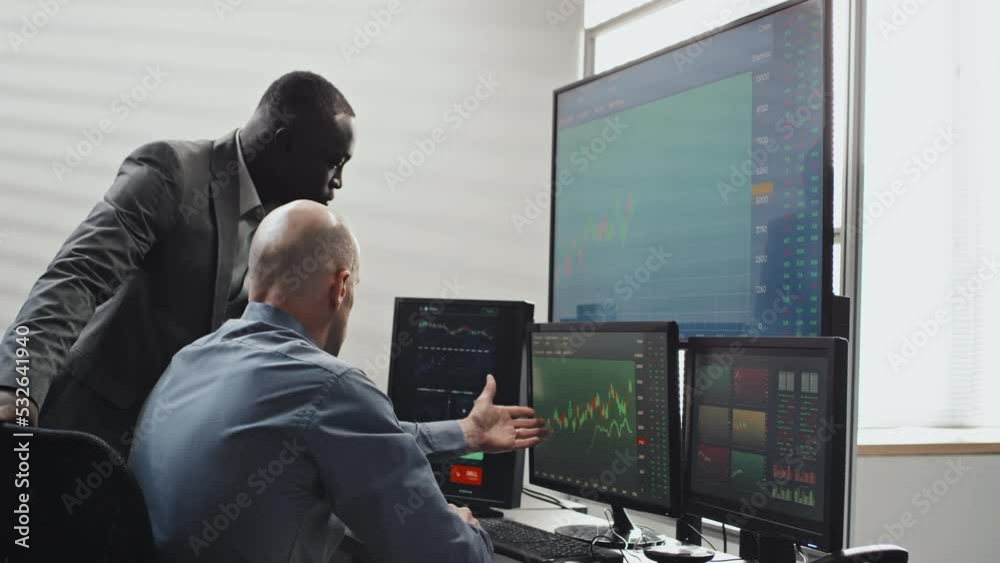 Mature Caucasian man and his young Black colleague looking at stock trading stats on computer monitors and discussing volatility Mature Caucasian man and his young Black colleague looking at stock trading stats on computer monitors and discussing volatility