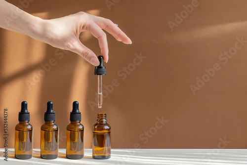 Woman's hand holds pipette with serum over an open bottle on light brown background, in rays of sunlight. Unmarked containers with cosmetics close-up. Open, closed bottles with oil. Concept of mockup