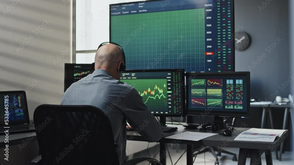 Professional brokerage worker holding document sitting in front of computer monitors checking currency stats Professional brokerage worker holding document sitting in front of computer monitors checking currency stats