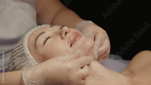Young hypnotic face woman receiving anti-ageing facial massage in spa salon relax. Wellness body skin care face beauty treatment, rejuvenation procedure getting head face massage. Black background