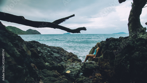 Sexy hot woman meditates, relaxes on rock crack reef hill in stormy morning cloudy sea. Girl in blue swimsuit. Concept feminine, sexual vaginal health, hygiene, womanly, freedom, freshness, recovery