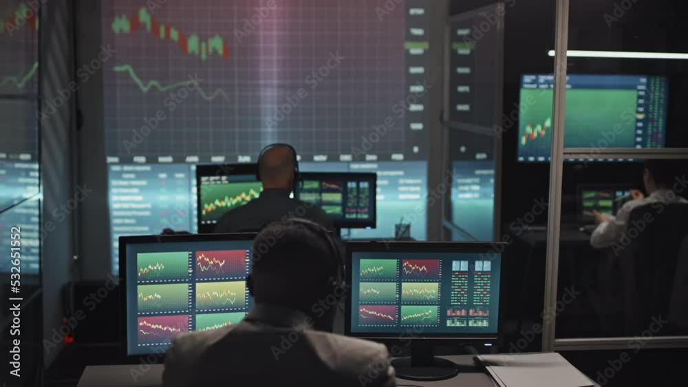 Group of unrecognizable brokerage specialists sitting at desks in office late in evening working with currency trading charts on computer screens Group of unrecognizable brokerage specialists sitting at desks in office late in evening working with currency trading charts on computer screens