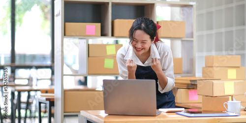 Startup small business entrepreneur SME or freelance Asian woman using a laptop with box, Young success Asian woman with her hand lift up, online marketing packaging box and delivery, SME concept.