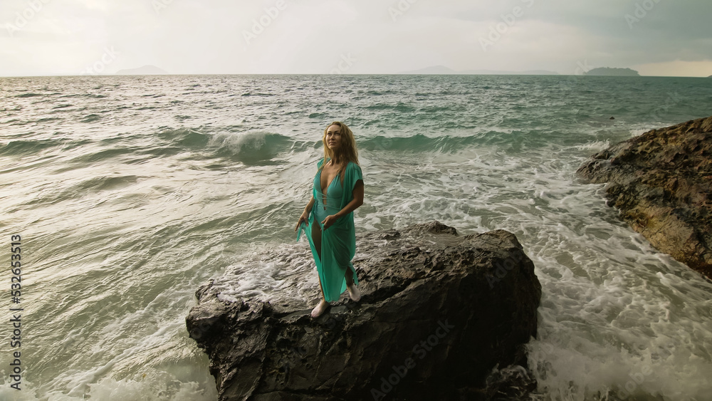 Seductive long-haired woman walks on rock of sea reef stone, stormy cloudy ocean. Woman in blue swimsuit dress tunic. Concept rest in sea, tropical resort coastline traveling tourism summer holidays