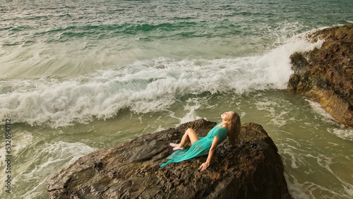 Seductive long-haired woman sitting on rock of sea reef stone, stormy cloudy ocean. Woman in blue swimsuit dress tunic. Concept rest in sea, tropical resort coastline traveling tourism summer holidays