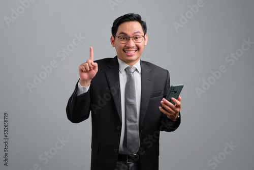 Portrait of cheerful handsome young businessman in formal suit using smartphone having an idea and pointing upwards isolated on grey background