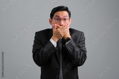Shocked handsome young businessman in formal suit covering mouth with hands isolated on grey background