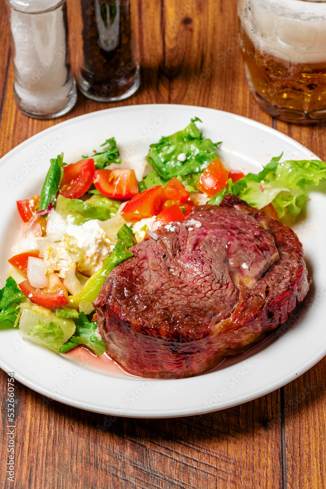 Cooked thin juicy beef steak on a white plate with green salad on a wooden table. Premium high quality meat dinner. Soft and tender meat cut prepared to highest standard.