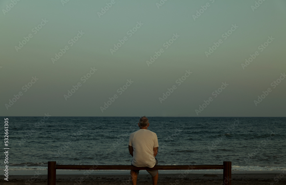Adult man sitting on wooden fence looking at view of sea Adult man sitting on wooden fence looking at view of sea