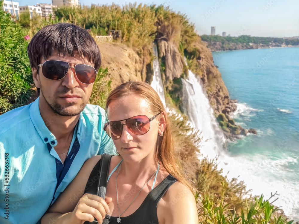 Adult couple taking a selfie in front of Lower Duden Waterfall in Antalya, Turkey. Portrait of a young man and woman in sunglasses against a blurred seascape Adult couple taking a selfie in front of Lower Duden Waterfall in Antalya, Turkey. Portrait of a young man and woman in sunglasses against a blurred seascape