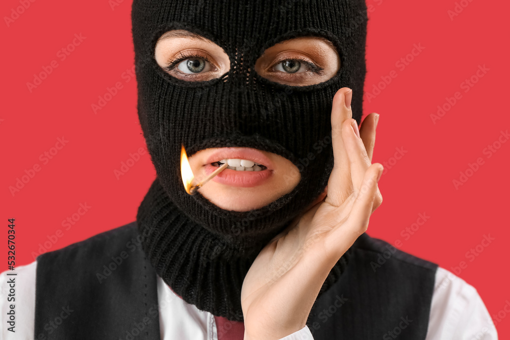 Portrait of young woman in balaclava with burning match in mouth on red background Portrait of young woman in balaclava with burning match in mouth on red background