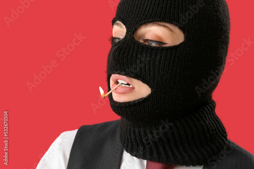 Portrait of young woman in balaclava with burning match in mouth on red background © Pixel-Shot
