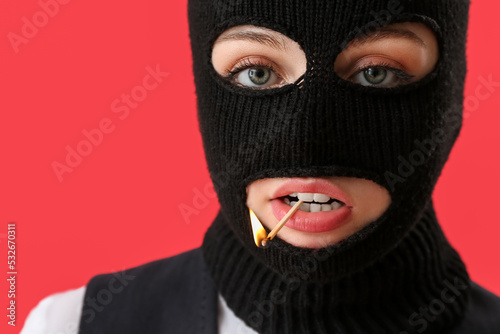 Portrait of young woman in balaclava with burning match in mouth on red background