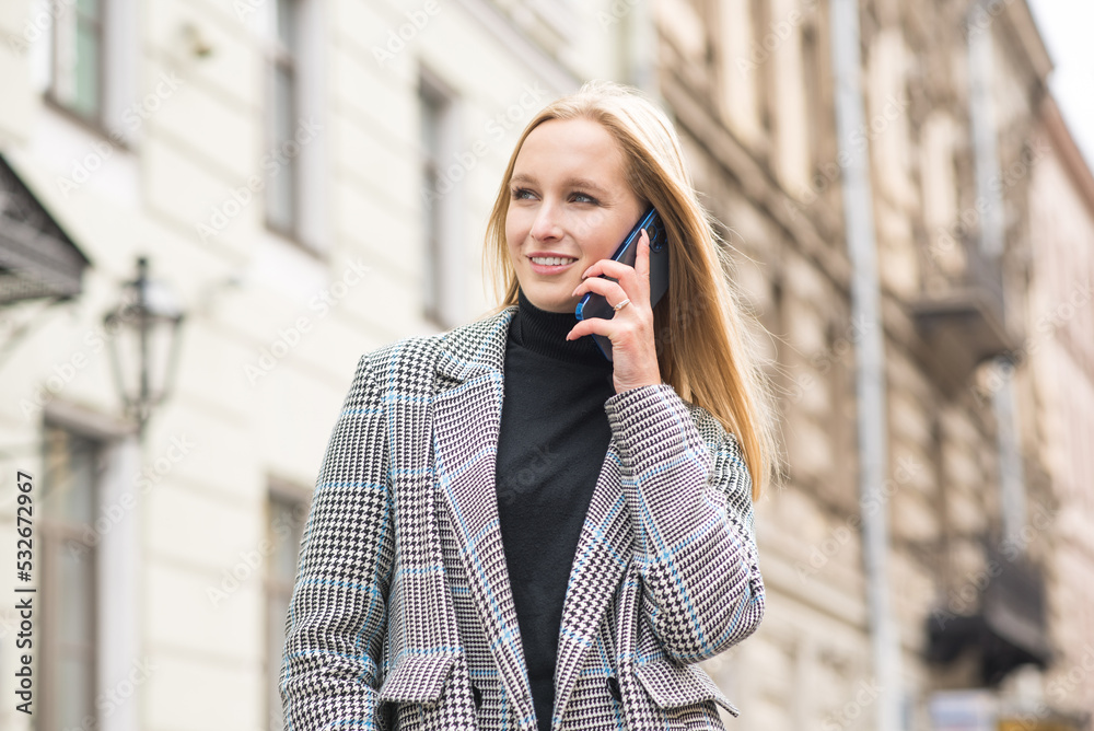 A young Caucasian girl in a coat is talking on a mobile phone and attentively listens to information. A businesswoman works walking in the city on an autumn day. The girl uses a smartphone and smiles. A young Caucasian girl in a coat is talking on a mobile phone and attentively listens to information. A businesswoman works walking in the city on an autumn day. The girl uses a smartphone and smiles.