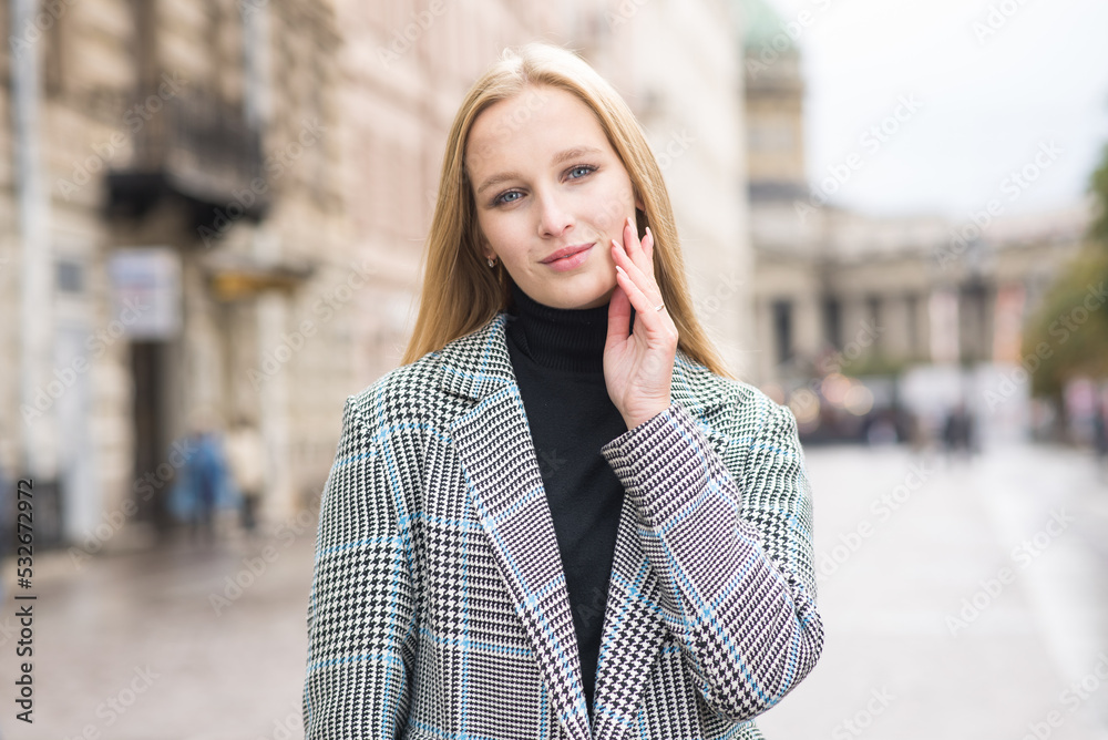 Fototapeta premium Portrait of a young and elegant Caucasian blonde girl in autumn in the city. The concept of femininity and style. A pretty girl is happy to pose in the city on the street in a coat.