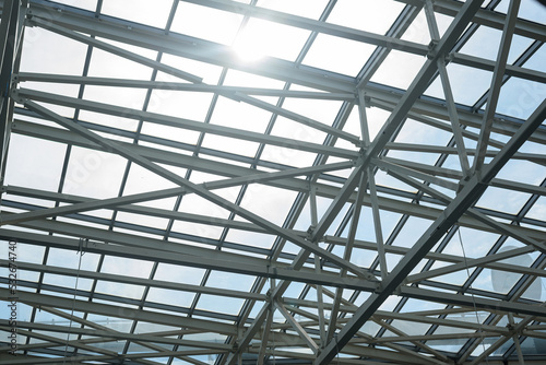 Sunlight breaks through abstract metal frame and blue glass construction. Bright sun illuminates modern structural roof facade of mall