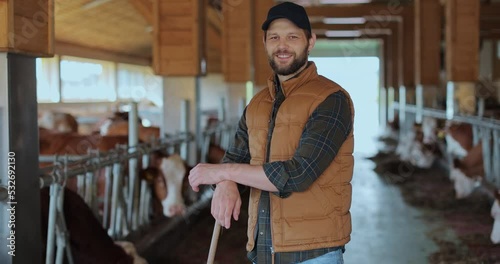 Portrait of handsome man farm at cowshed feedlots. Young man with beard farmer cleaning cowshed barn by pork. Cows eating grass on background. photo