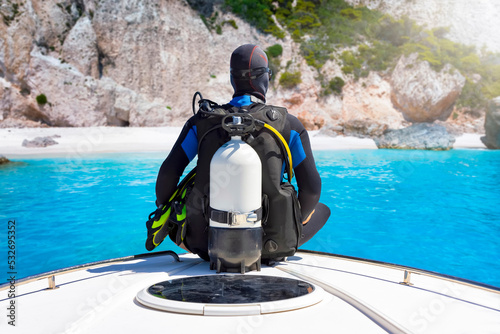 A scuba diver with his equipment sits on a bow of a boat in front of a beautiful beach with turquoise sea photo