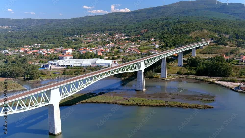 railway bridge over the ulla river, the village and industrial buildings and the forested mountains in the sunny blue sky orizonte, drone shot traveling diagonal right, Catoira, Galicia, Spain railway bridge over the ulla river, the village and industrial buildings and the forested mountains in the sunny blue sky orizonte, drone shot traveling diagonal right, Catoira, Galicia, Spain