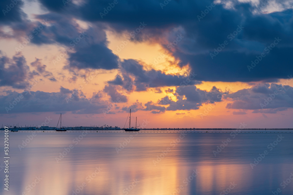 Sunrise over the Mediterranean sea, on a cloudy summers morning.  The sun is reflecting on the calm sea, on which there are a few sailing boats
