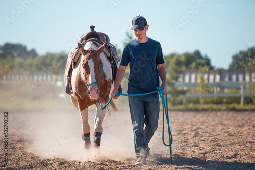 A young male cowboy leads his pinto horse through a sandy dusty arena. Western horse training on rope and rope halter. Horsemenship. Summer photo of a saddled western horse and a man in the manege. photo