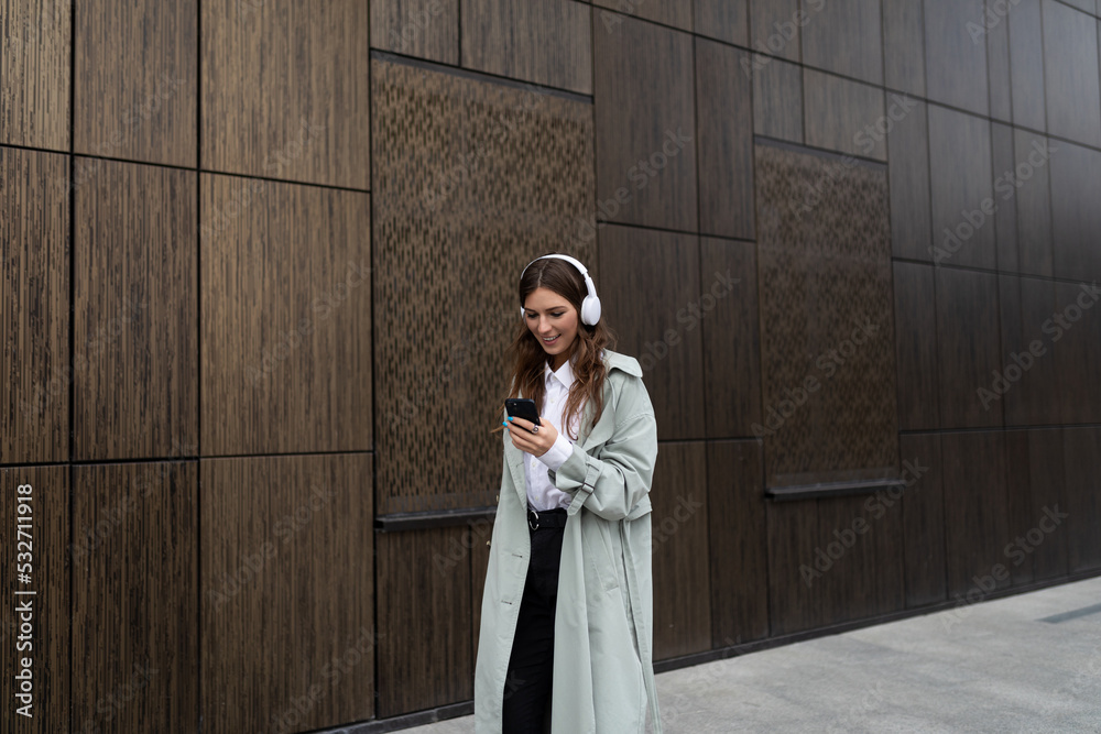 a young woman walks around the city against the background of the brown walls of a modern building in headphones listening to music a young woman walks around the city against the background of the brown walls of a modern building in headphones listening to music