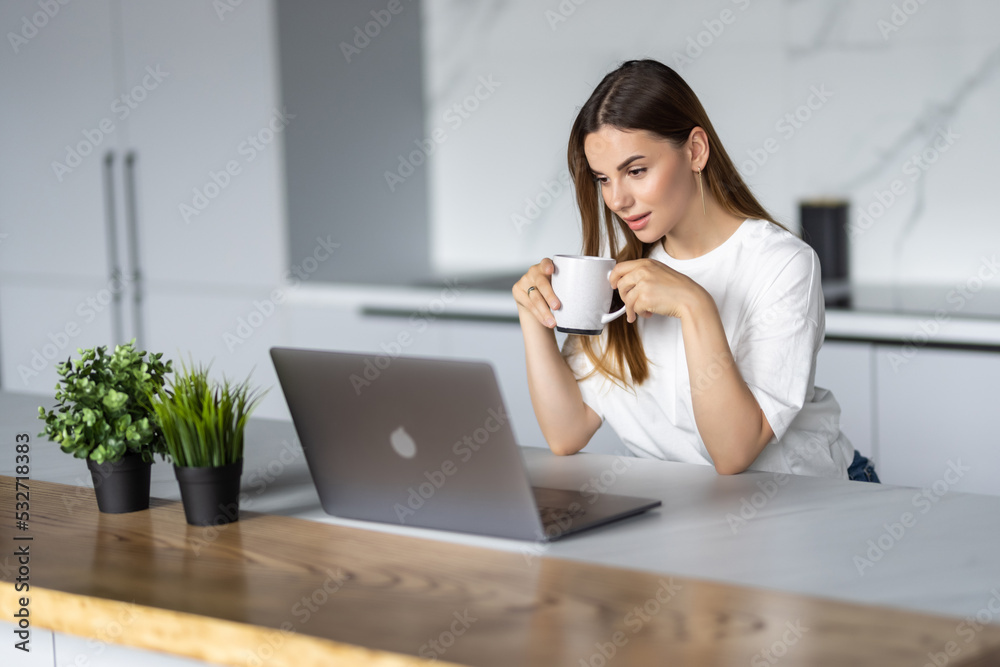 Young woman drinking morning coffee look laptop while sitting in the kitchen at home Young woman drinking morning coffee look laptop while sitting in the kitchen at home