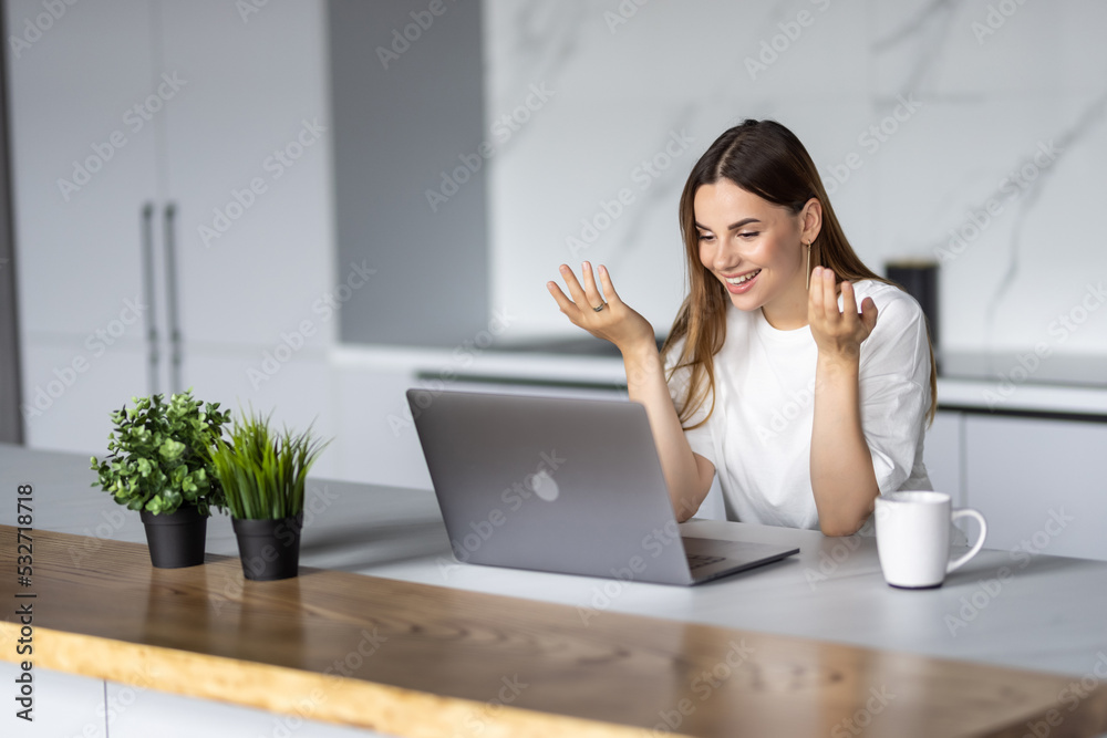 Young pretty woman talking on video call and waving hand while sitting at table inthe kitchen Young pretty woman talking on video call and waving hand while sitting at table inthe kitchen