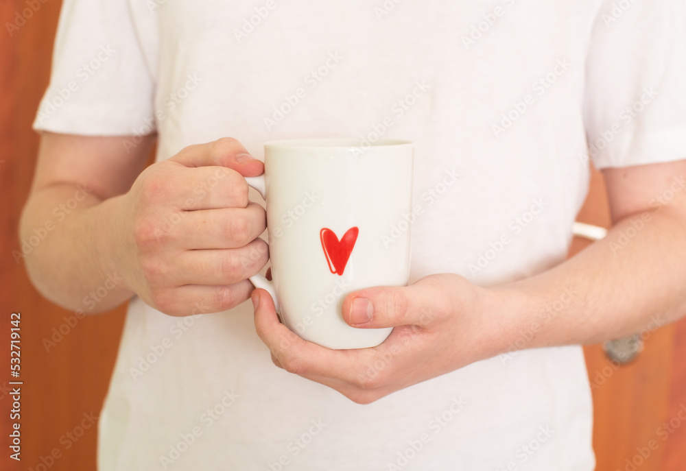 The hands of a young man in a white T-shirt hold a white cup with a red heart pattern. Tea time. Coffee time. Life style