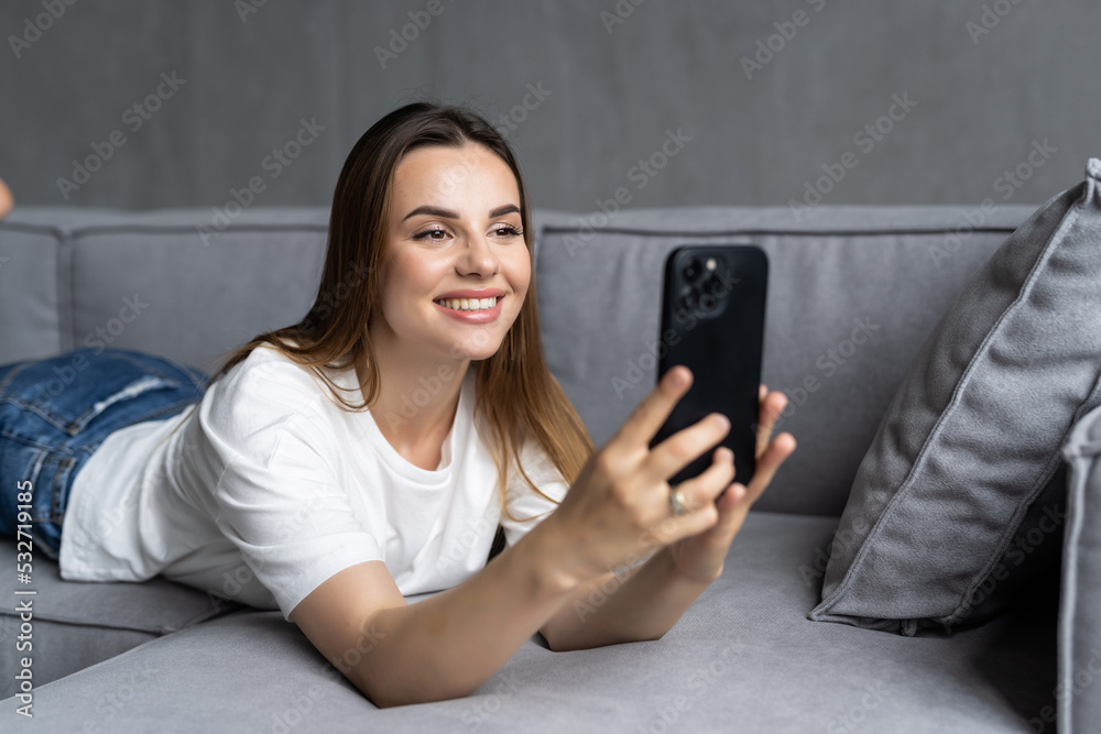 Attractive young woman smiling as she reads an sms message on her mobile while relaxing barefoot on a sofa in her living room Attractive young woman smiling as she reads an sms message on her mobile while relaxing barefoot on a sofa in her living room