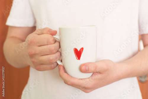 The hands of a young man in a white T-shirt hold a white cup with a red heart pattern. Tea time. Coffee time. Life style